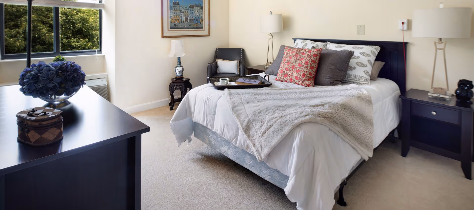 Well-lit furnished bedroom with a made bed, nightstands, lamps, a dresser, and a window showing greenery.