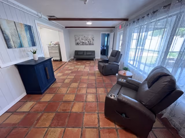 Sunlit common lounge with terra-cotta tile floor, two reclining chairs by sheer-curtained windows, a small sofa against the far wall, and a blue cabinet.