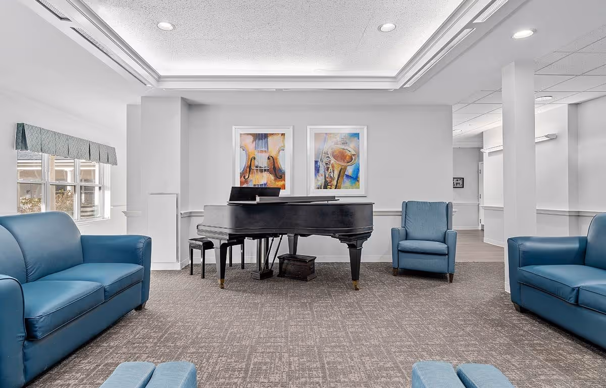 A bright and spacious lounge area featuring a black grand piano at the center, flanked by two blue armchairs and two blue sofas. The room has a large window with a patterned valance, two colorful framed paintings of musical instruments on the wall behind the piano, and a carpeted floor.