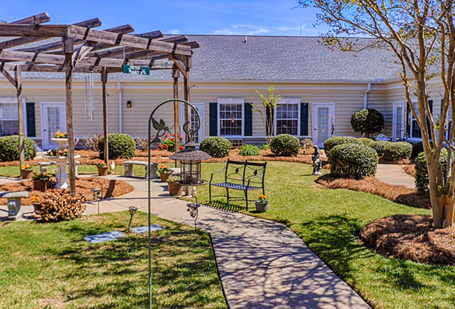 Outdoor garden area at Sumter Grove Senior Living with a paved walkway, a wooden pergola, a metal bench, various potted plants, trimmed bushes, and a building with white siding and green shutters in the background under a clear blue sky.