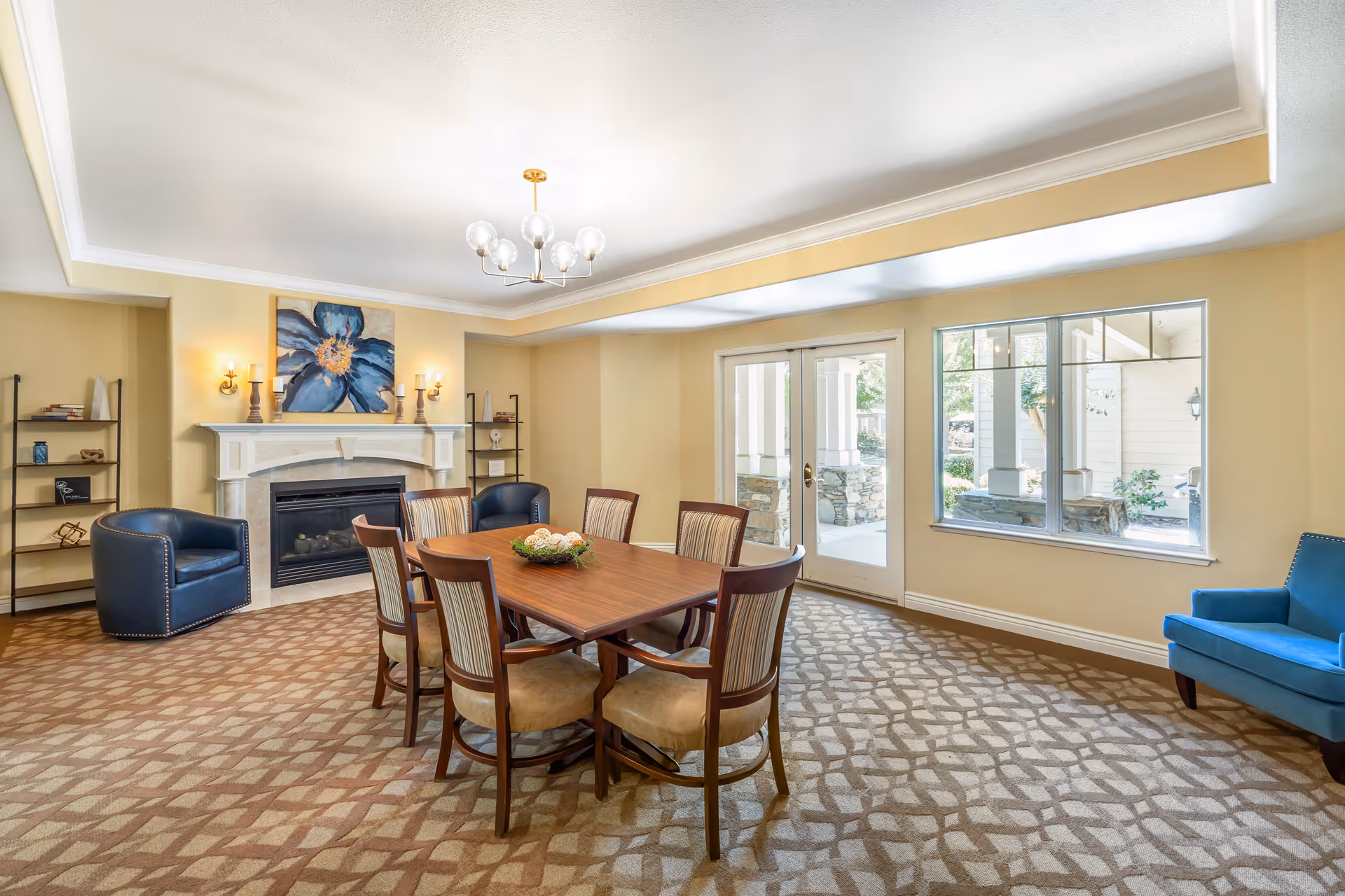 Bright communal dining room with a wooden table and six chairs, a fireplace, armchairs, and large windows and doors to the outside.