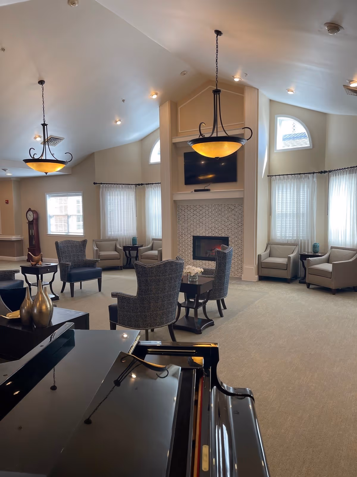 Bright communal lounge with armchairs around a fireplace and TV, chandeliers overhead, and a grand piano in the foreground.