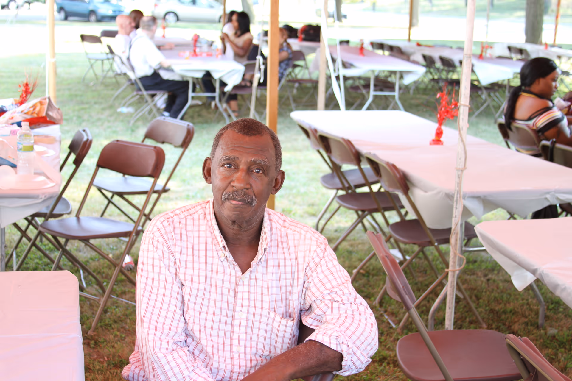 An elderly man wearing a checkered shirt sits at a table under a large outdoor tent with other people seated at tables in the background. The tables are covered with white tablecloths and there are folding chairs around them. The setting appears to be a social gathering or event on a grassy area.
