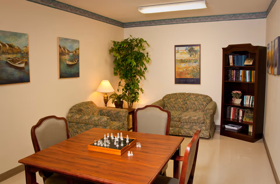 A cozy common area with a wooden table and four chairs, a chess set on the table, two patterned armchairs and a loveseat, a floor lamp, a tall green plant, a bookshelf filled with books and a small potted plant, and several framed paintings on the walls.