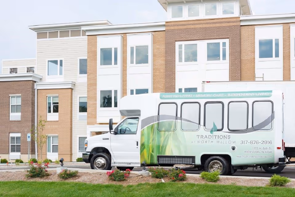 A white shuttle bus with green leaf graphics parked in front of a multi-story brick and siding building. The bus has signage for Traditions at North Willow, including contact information and services like assisted living, memory care, and independent living. The building has multiple windows and a landscaped area with grass and small bushes in front.