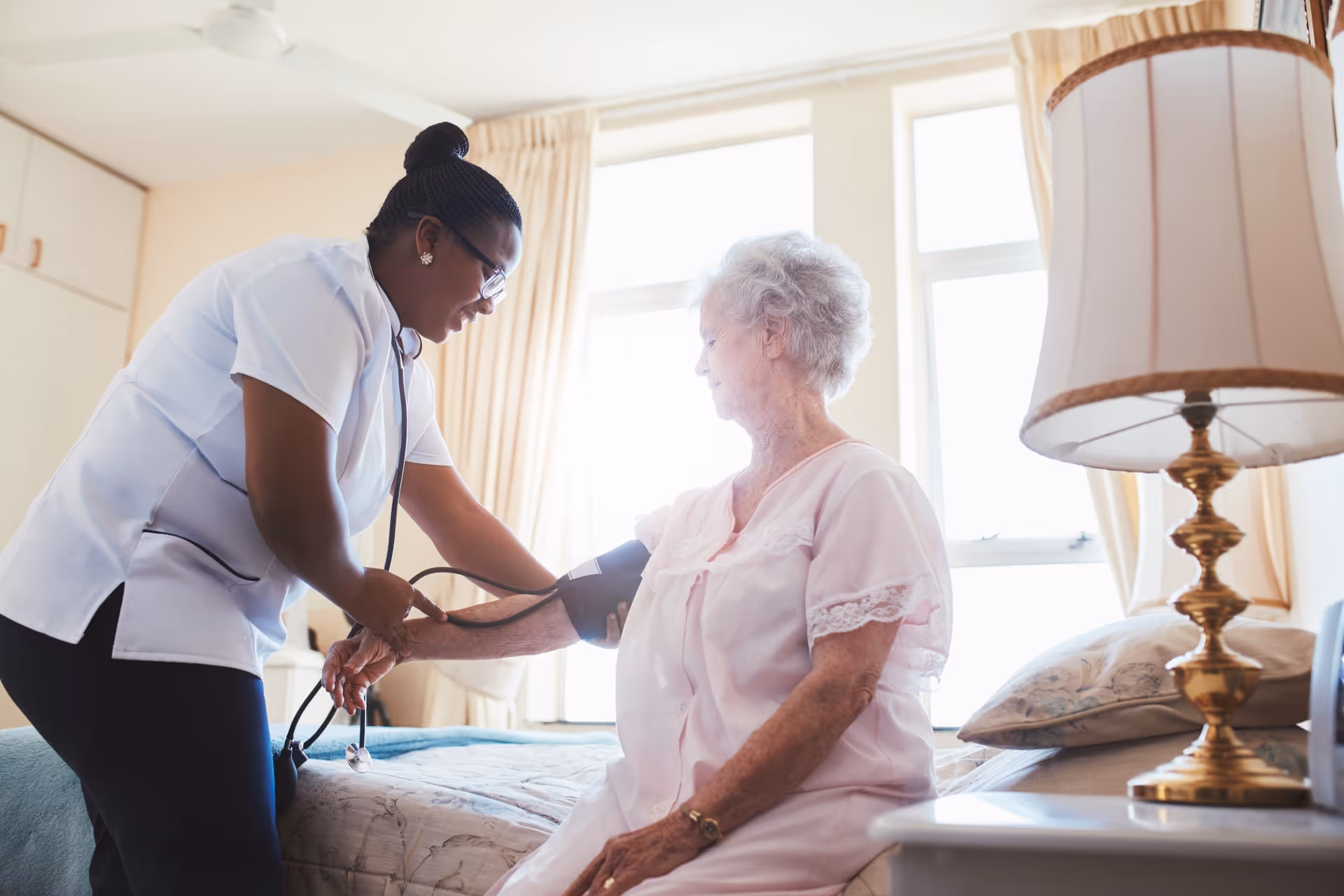 A caregiver measures an elderly woman's blood pressure while she sits on a bed in a bright bedroom.