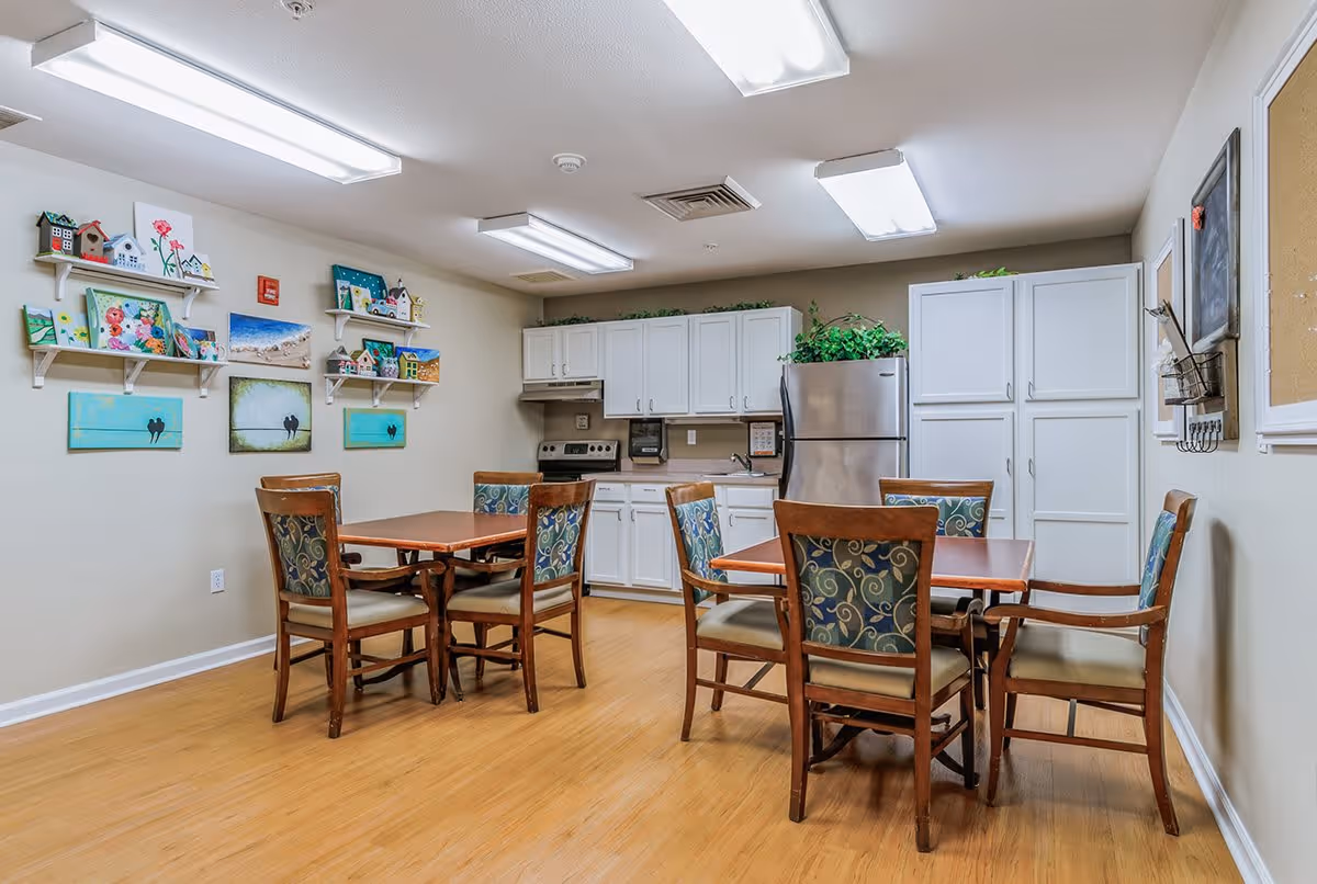 A bright kitchen and dining area with wooden floors, two wooden tables each surrounded by four chairs with patterned upholstery. The kitchen has white cabinets, a stainless steel refrigerator, and a stove. The walls are decorated with colorful paintings and small decorative birdhouses on white shelves.