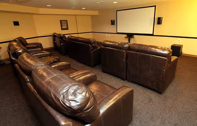 A small theater room with multiple rows of brown leather recliner chairs facing a large white projection screen mounted on a beige wall. The room has carpeted flooring and recessed ceiling lights.