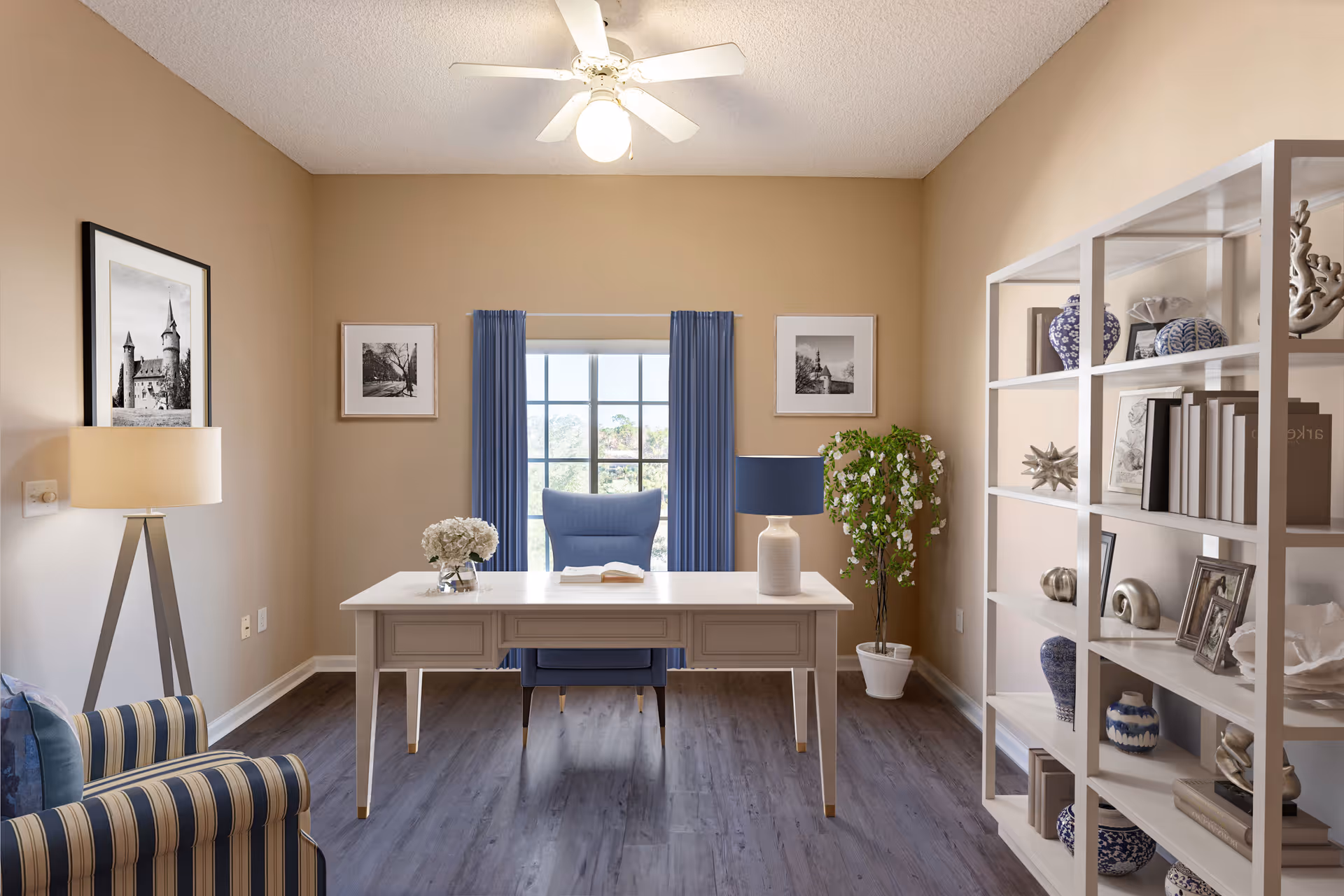 A bright and neatly arranged office room with a white desk and a blue chair positioned in front of a window with blue curtains. The room features a striped armchair, a floor lamp, framed black and white pictures on the walls, a white shelving unit with decorative items and books, and a potted plant. The ceiling has a white fan with a light.