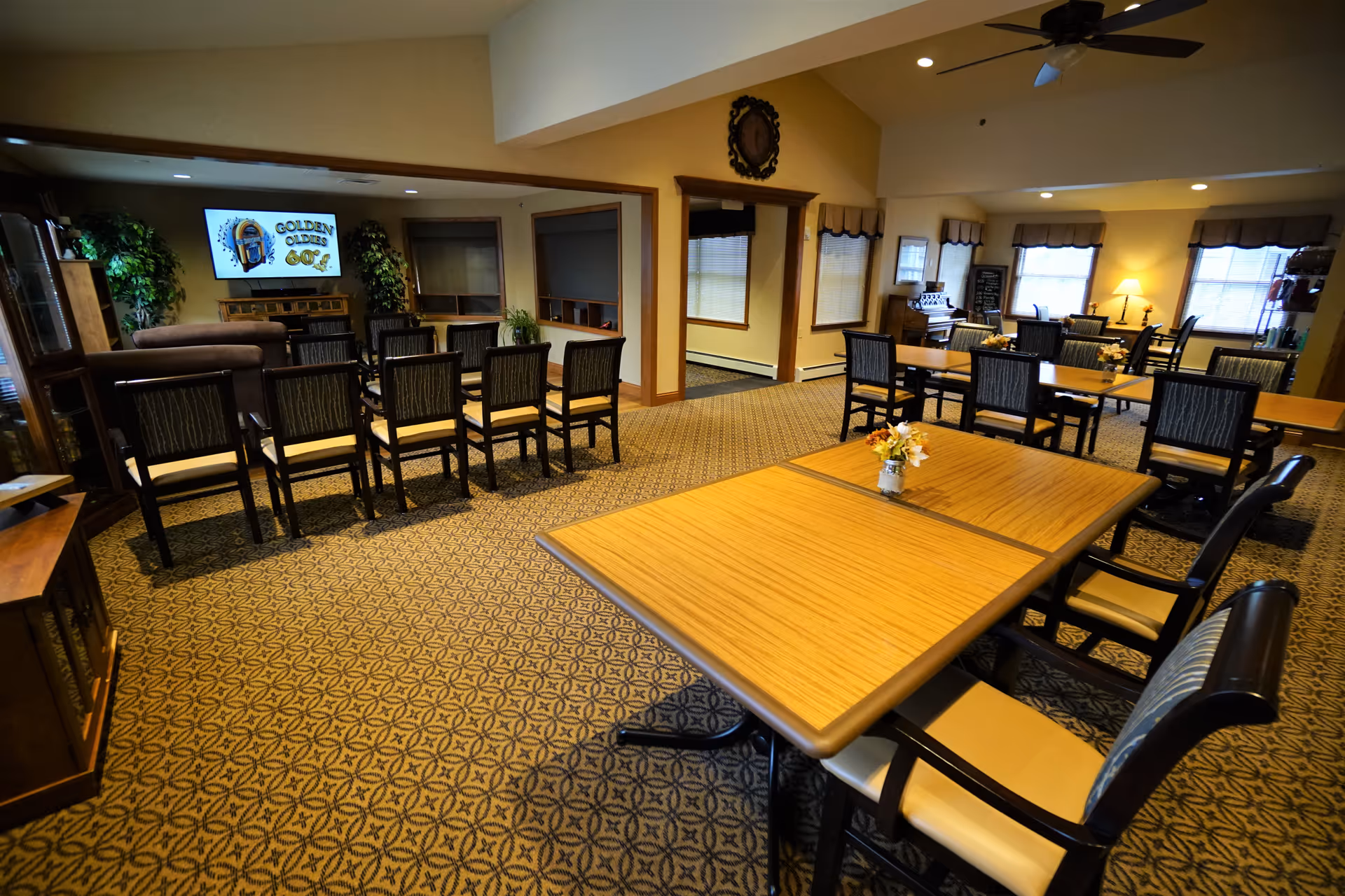 A spacious common area with multiple wooden tables and chairs arranged neatly on a patterned carpet. The room has warm lighting, several windows with valances, a ceiling fan, and a television mounted on the wall displaying a 'Golden Oldies' logo. There are plants and furniture pieces along the walls, creating a cozy and inviting atmosphere.