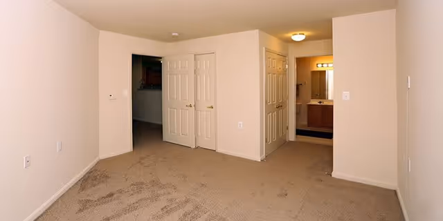 Empty room with beige carpet and cream-colored walls, featuring two closed double doors and an open doorway leading to a bathroom with a sink and mirror visible.