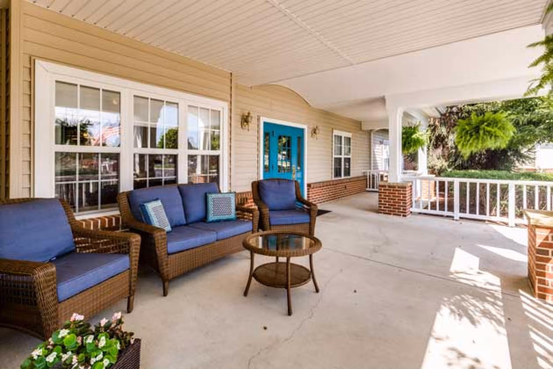 Covered outdoor porch area with wicker furniture including a loveseat and two chairs with blue cushions, a round glass-top coffee table, hanging green plants, beige siding, white framed windows, and a bright blue door.