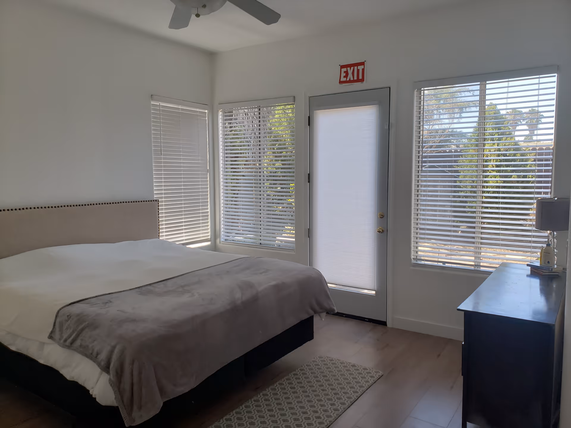 A bright bedroom with a large bed covered with white and gray bedding. The room has three windows with white blinds and a door with a white shade and an exit sign above it. There is a dark wooden dresser with a lamp and some items on top. The floor is wooden with a small patterned rug near the bed.