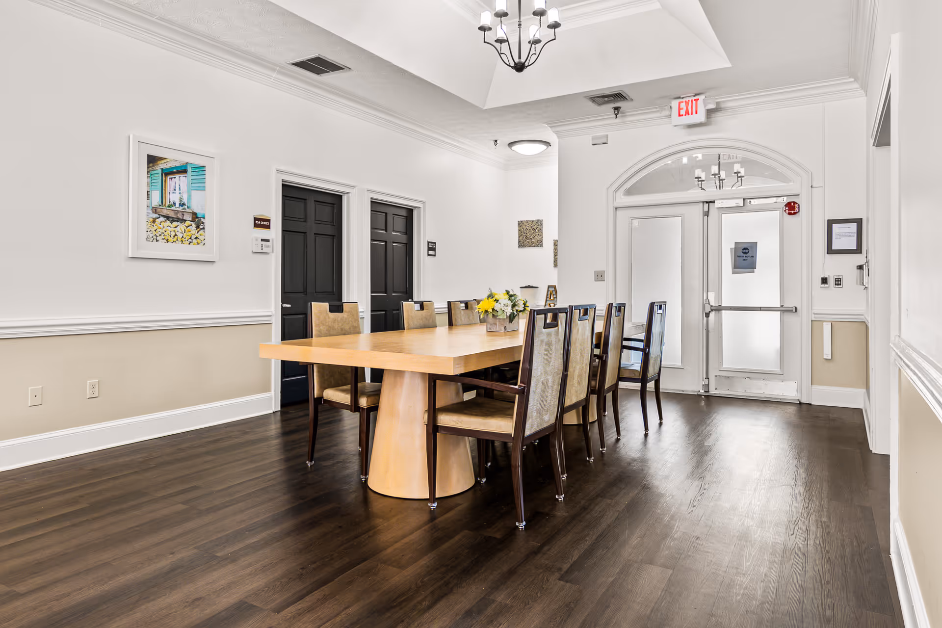 A bright dining-style room with a long wooden table surrounded by chairs and double glass exit doors.