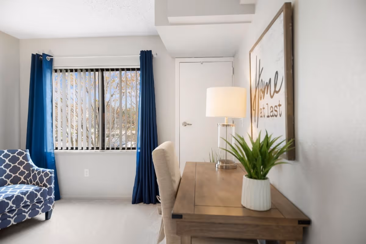 A bright room with a wooden desk holding a lamp and a potted plant. A beige upholstered chair is positioned at the desk. On the wall above the desk is a framed sign that reads 'Home at last'. To the left, there is a window with vertical blinds and blue curtains, and a blue patterned armchair is partially visible.
