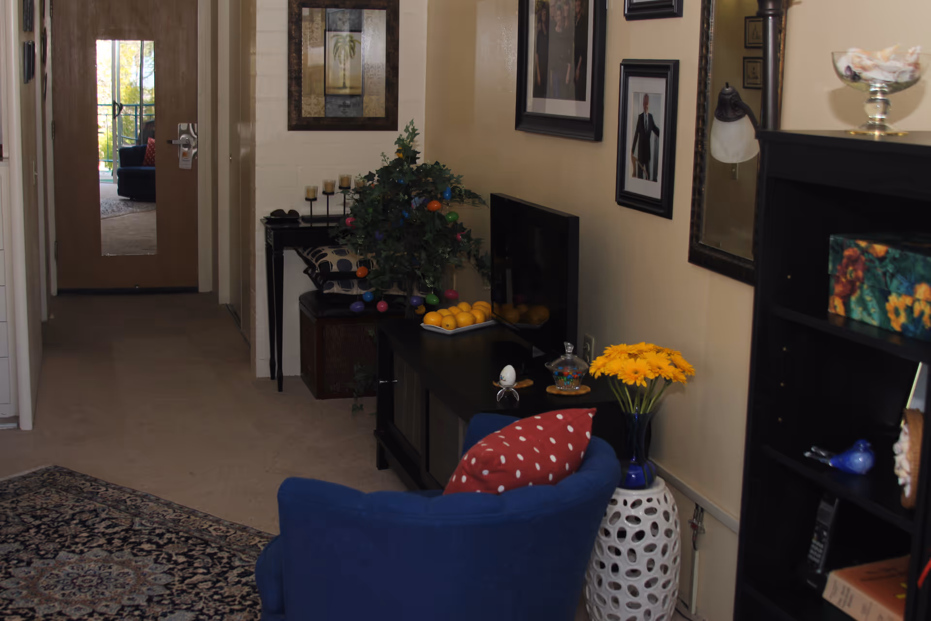 Cozy living room area with a blue armchair, TV console, bookshelf and decorative flowers visible down a hallway.