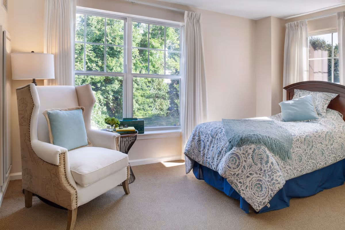 A bright bedroom with a large window showing green trees outside. The room features a comfortable armchair with a light blue cushion, a small side table with books and a plant, and a bed with a patterned blue and white bedspread and matching pillows. Light beige walls and carpet complete the cozy setting.