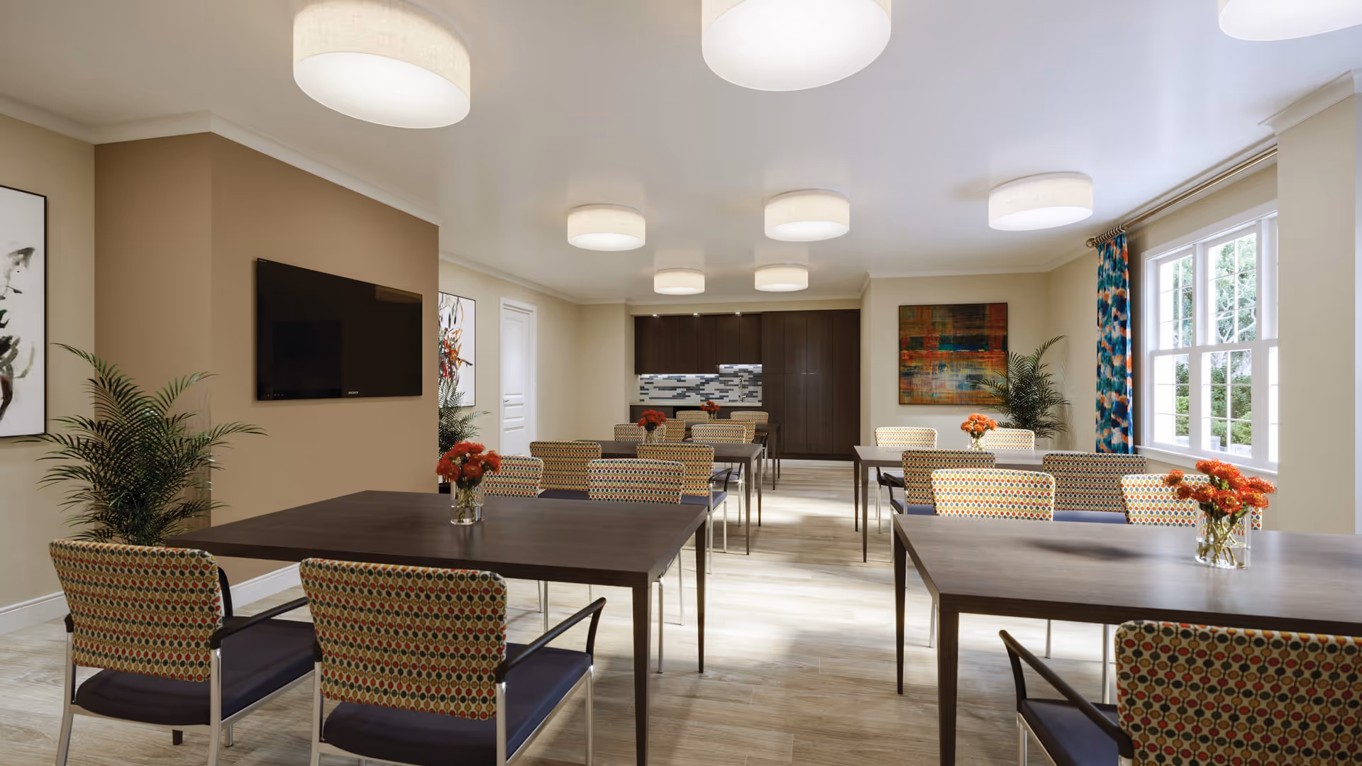 A bright dining room with multiple dark wood tables and patterned chairs. Each table has a small vase with orange flowers. The room features large windows with colorful curtains, modern ceiling lights, a wall-mounted TV, and decorative plants and artwork on the walls.