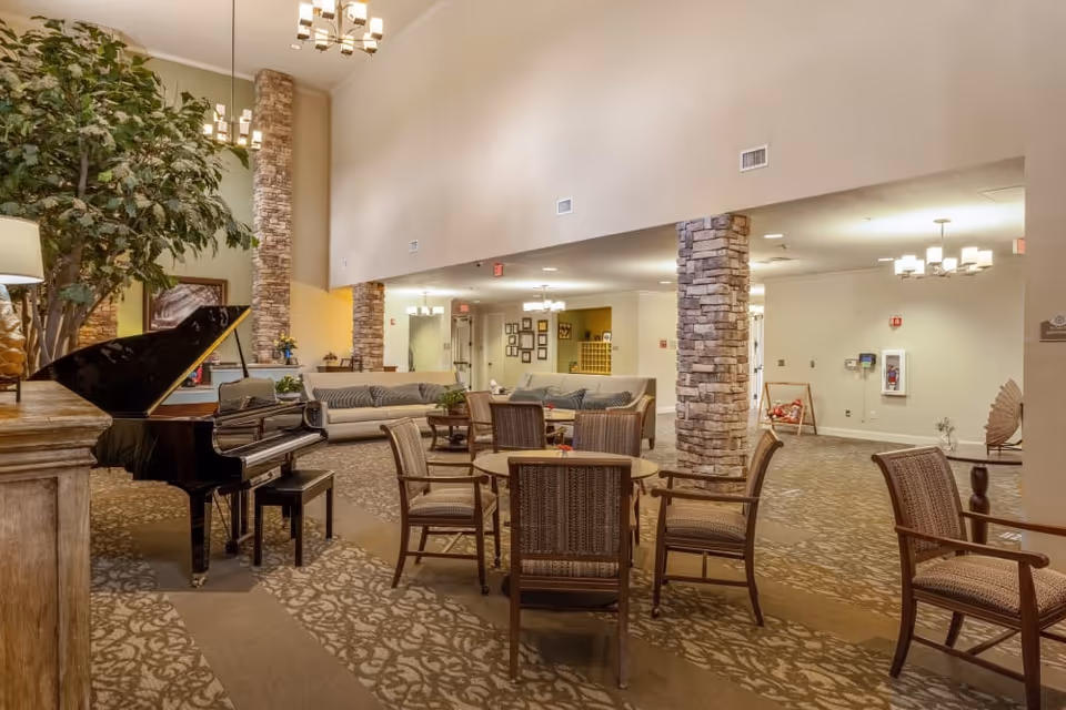 Spacious senior living lounge with a grand piano, round tables and chairs, sofas, stone columns, and hanging chandeliers.