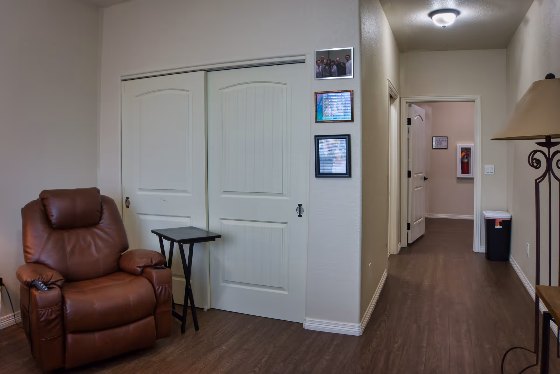 Interior view of a room with a brown leather recliner chair next to a small black side table. There are white double sliding closet doors on the wall behind the chair. The hallway extends to the right with a door open at the end, showing a fire extinguisher mounted on the wall. A lamp with a beige shade is partially visible on the right side of the image. The floor is dark wood, and the walls are painted light beige. Three framed pictures are hung on the wall near the closet doors.