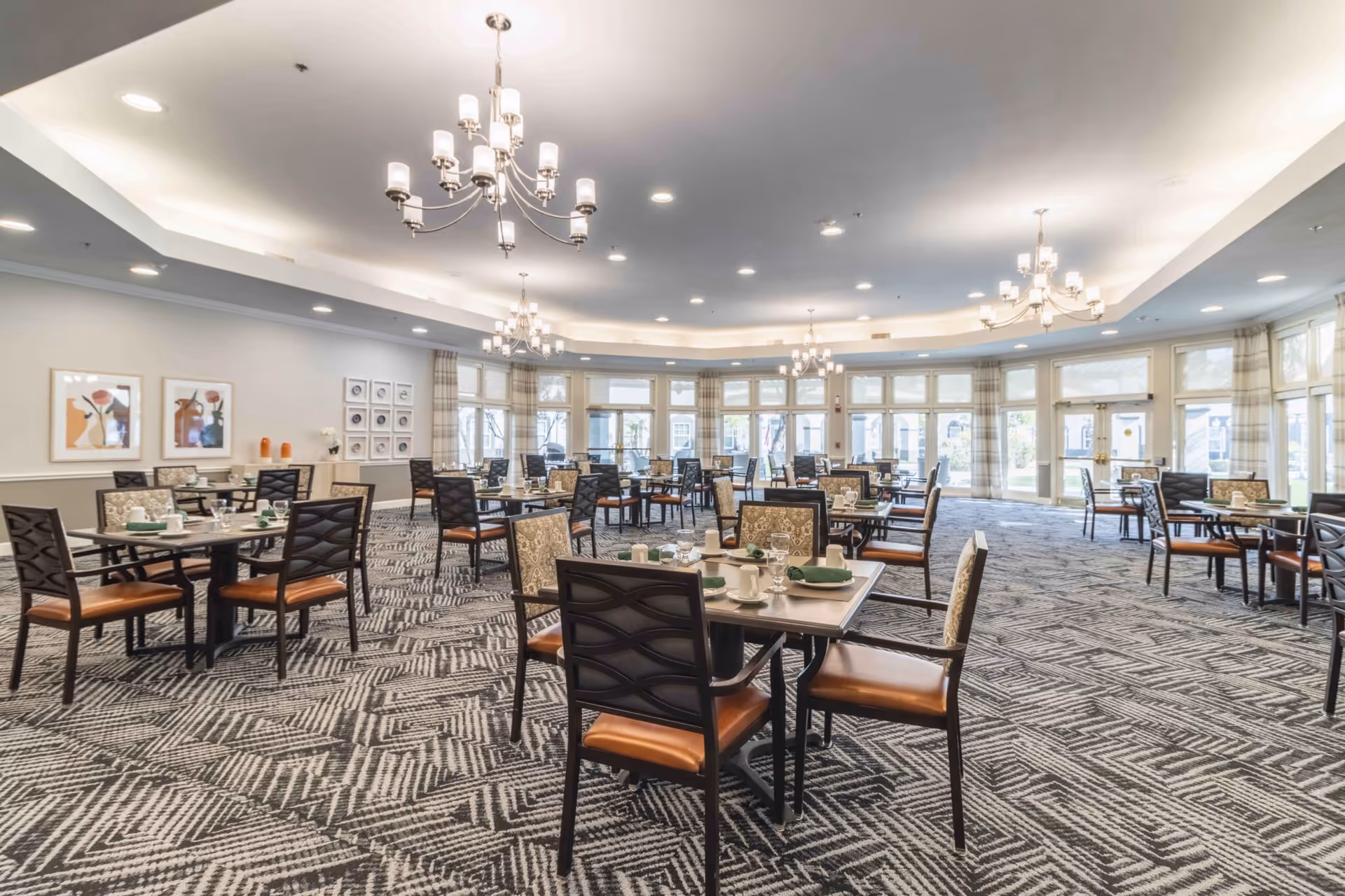 A spacious dining room with multiple tables and chairs arranged neatly. Each table is set with cups, glasses, and green napkins. The room features large windows allowing natural light to fill the space, modern chandeliers hanging from the ceiling, patterned carpet flooring, and framed artwork on the walls.