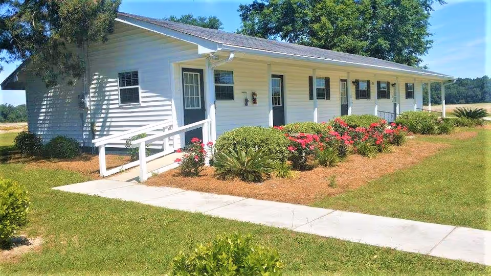 Single-story white building with multiple doors and windows, surrounded by a well-maintained garden with green bushes and pink flowers. A concrete walkway leads to the building, which has a ramp for accessibility. The sky is clear and blue.