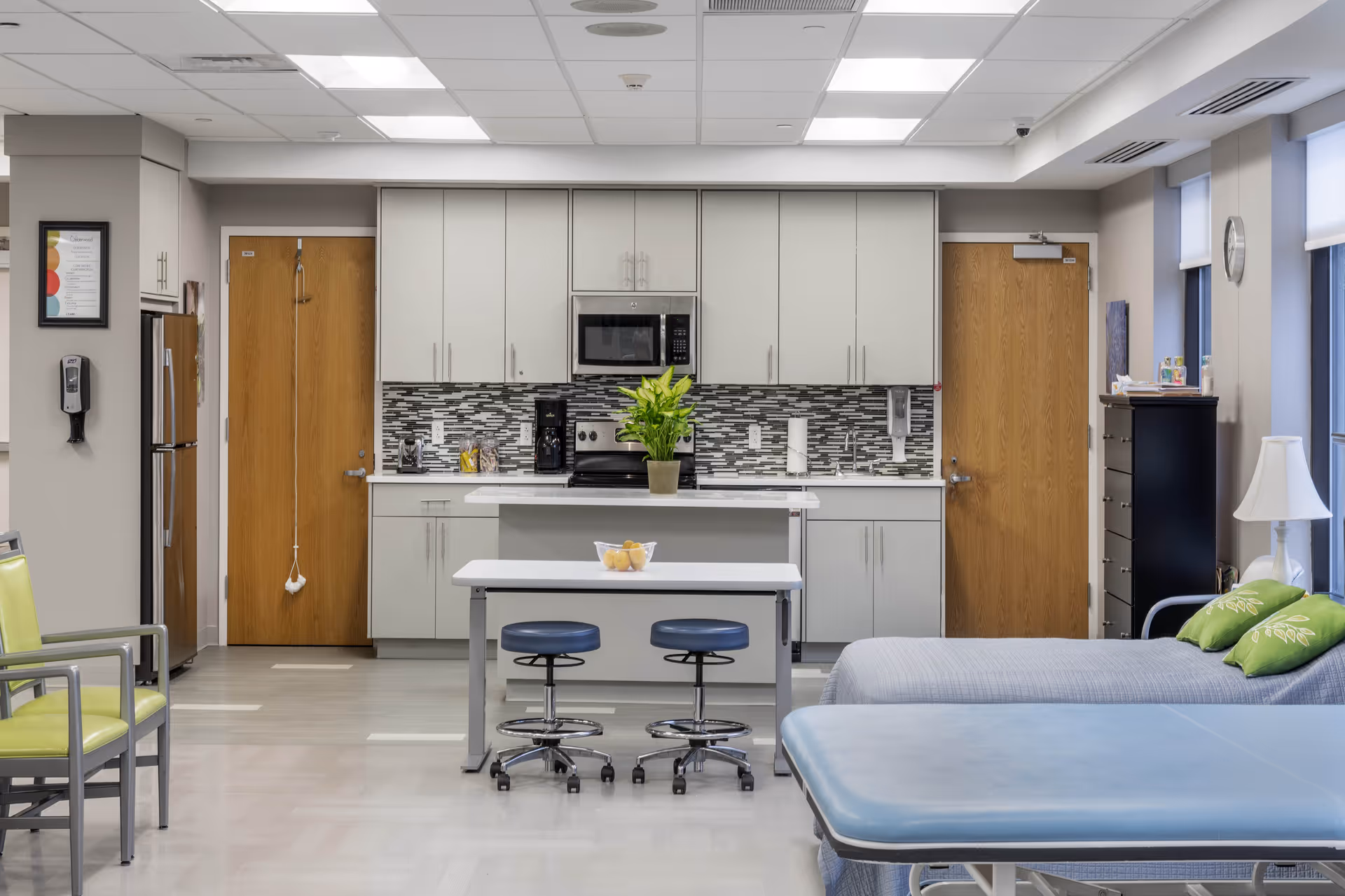 A bright, clean room featuring a kitchenette with white cabinets, a microwave, coffee maker, and a tiled backsplash. In front of the kitchenette is a small table with two blue stools and a bowl of lemons. To the right, there is a bed with green pillows and a blue cushioned bench or examination table. On the left, there are green chairs and a stainless steel refrigerator. The room has two wooden doors and large windows with blinds, letting in natural light.