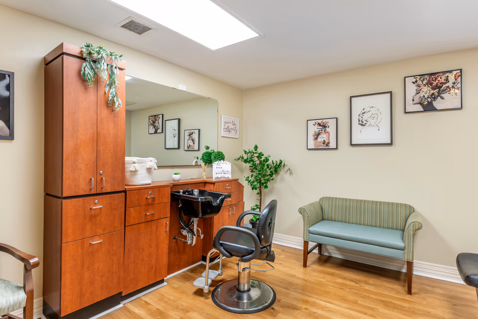 Small salon room with wooden cabinetry, a styling chair and sink, and a striped loveseat against a wall with framed artwork.