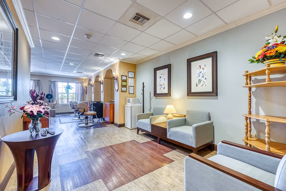 A bright and welcoming interior space with comfortable seating including armchairs and a small table with a lamp. The room features framed floral artwork on the walls, a wooden shelving unit with a colorful flower arrangement, and a small table with a vase of flowers near the entrance. In the background, there are salon-style chairs and large windows with curtains letting in natural light.