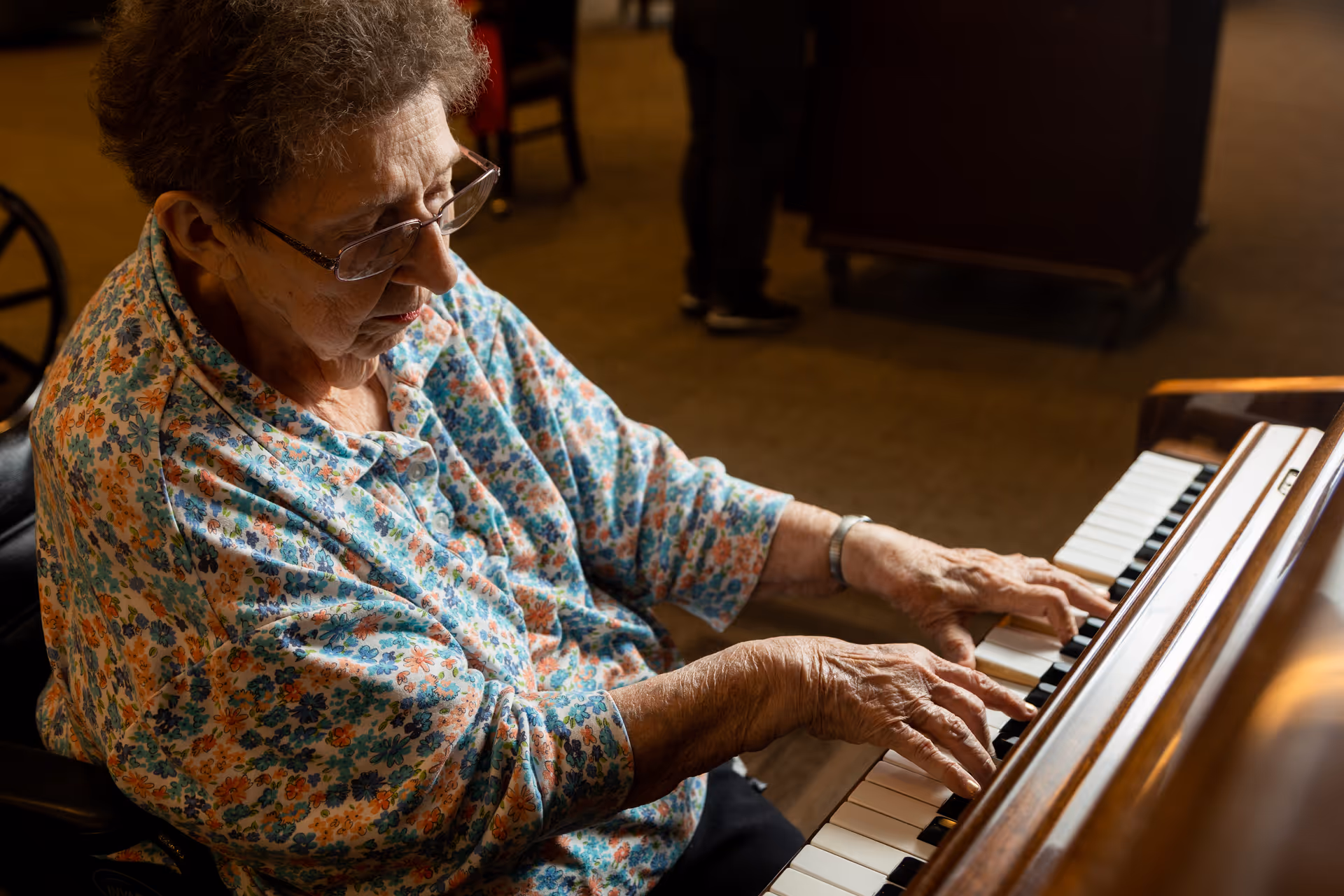 An elderly woman wearing glasses and a floral patterned shirt is playing a piano indoors, focusing on the keys with her hands positioned to play.