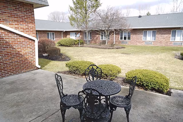 Outdoor patio area with a black metal table and four matching chairs on a concrete surface. Surrounding the patio is a grassy courtyard with trimmed bushes and a few leafless trees. In the background, there is a single-story brick building with multiple windows.