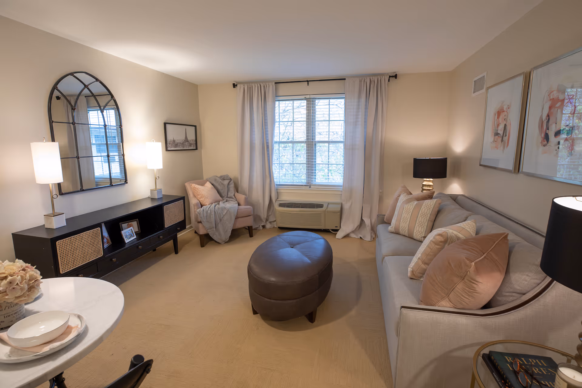 A cozy living room with a beige carpet, a light gray sofa adorned with multiple pillows, a round dark brown ottoman in the center, a pink armchair with a gray throw blanket, a black console table with two lamps and framed pictures, a large window with white curtains, and two framed abstract artworks on the wall.