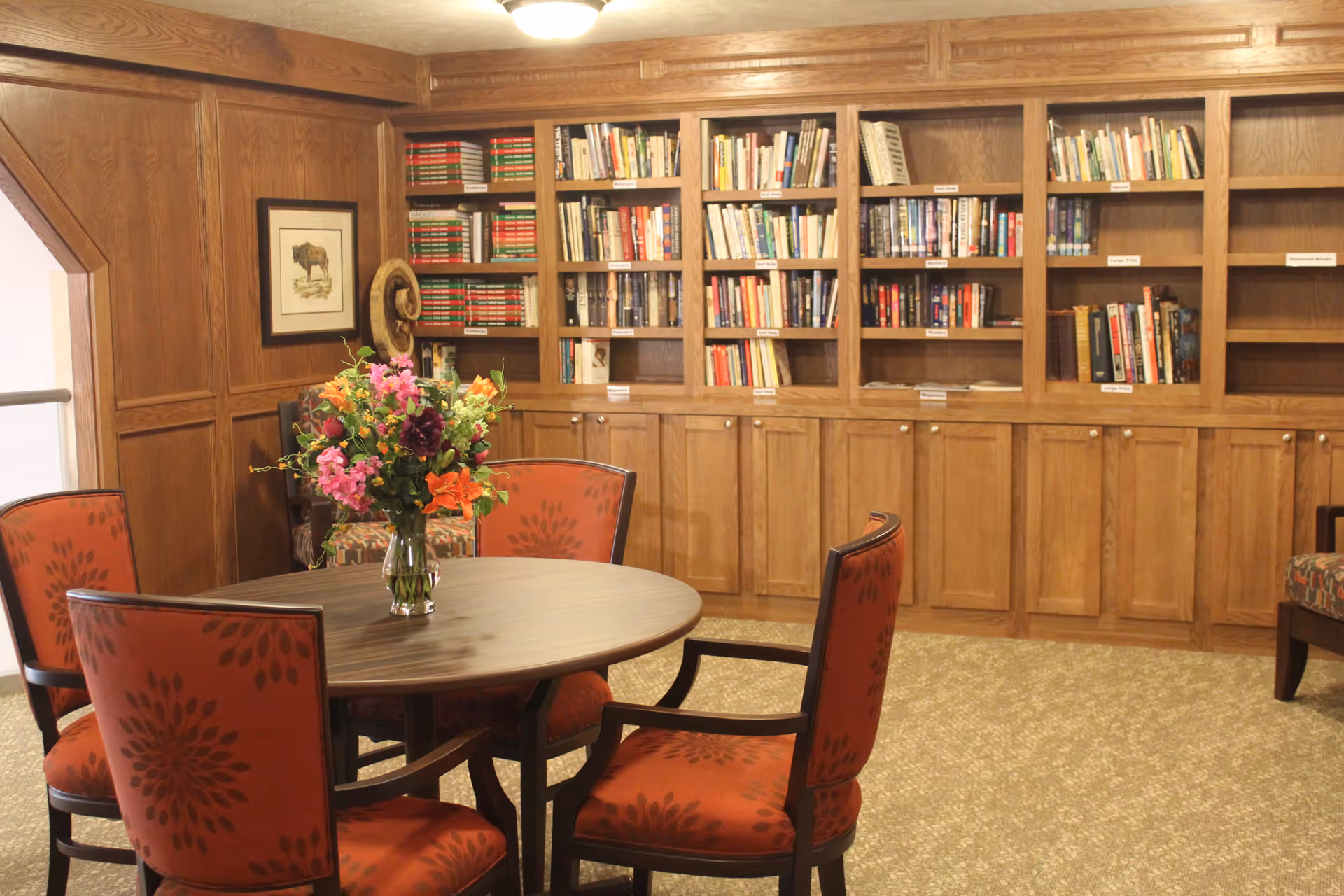 A cozy library or reading room with wooden bookshelves filled with books along the back wall. In the center, there is a round wooden table with a vase of colorful flowers on it, surrounded by four upholstered chairs with red patterned fabric. The room has wood-paneled walls and a beige carpeted floor.
