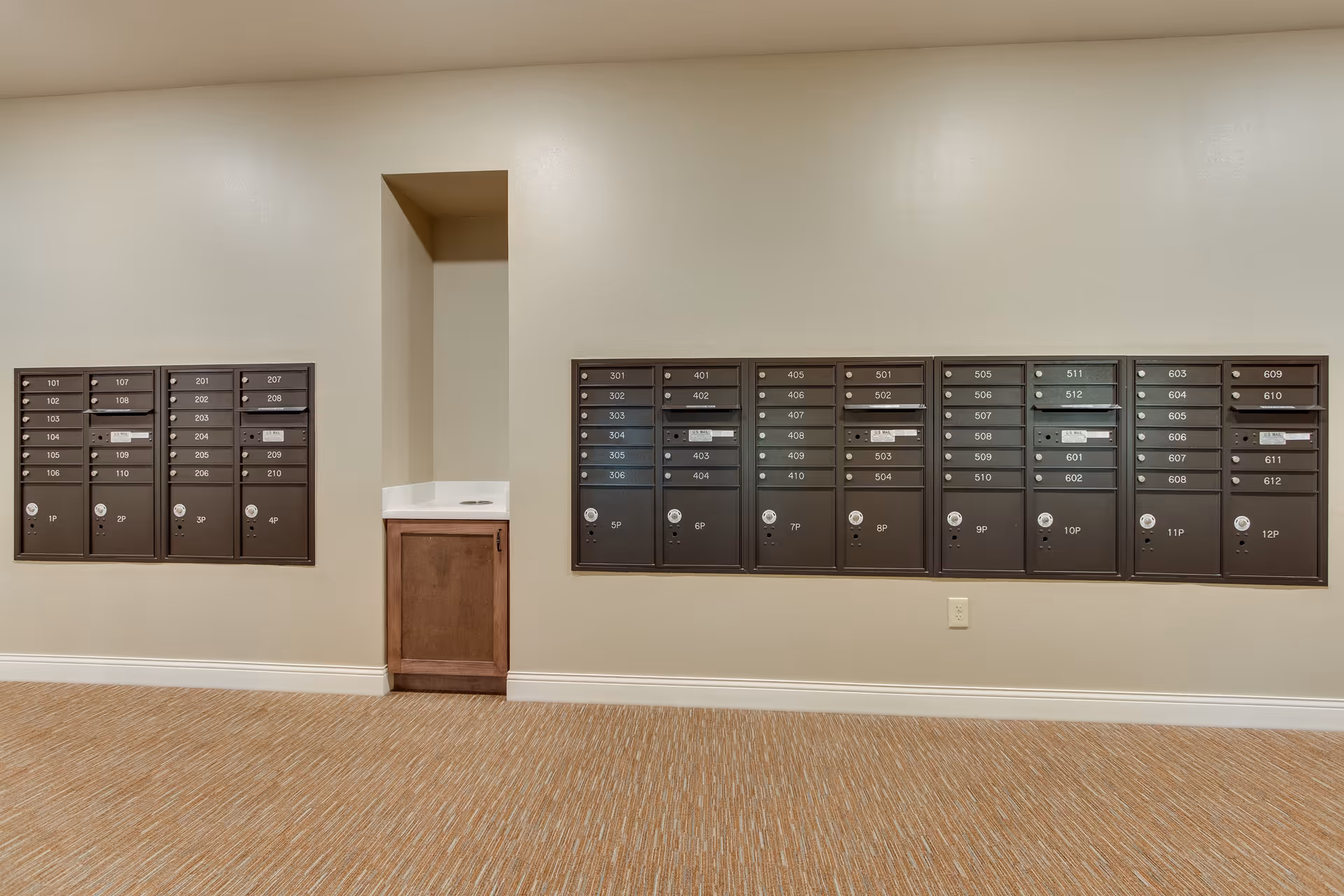 Interior view of a mailroom area with multiple black mailboxes mounted on a beige wall. The mailboxes are labeled with room numbers and arranged in two groups on either side of a small recessed area with a countertop and cabinet below. The floor is carpeted in a light brown pattern.