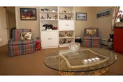 A cozy living room with two plaid armchairs facing a glass coffee table. Behind the chairs is a white built-in shelving unit with decorative items and framed pictures on the walls. The room has a warm, inviting atmosphere.