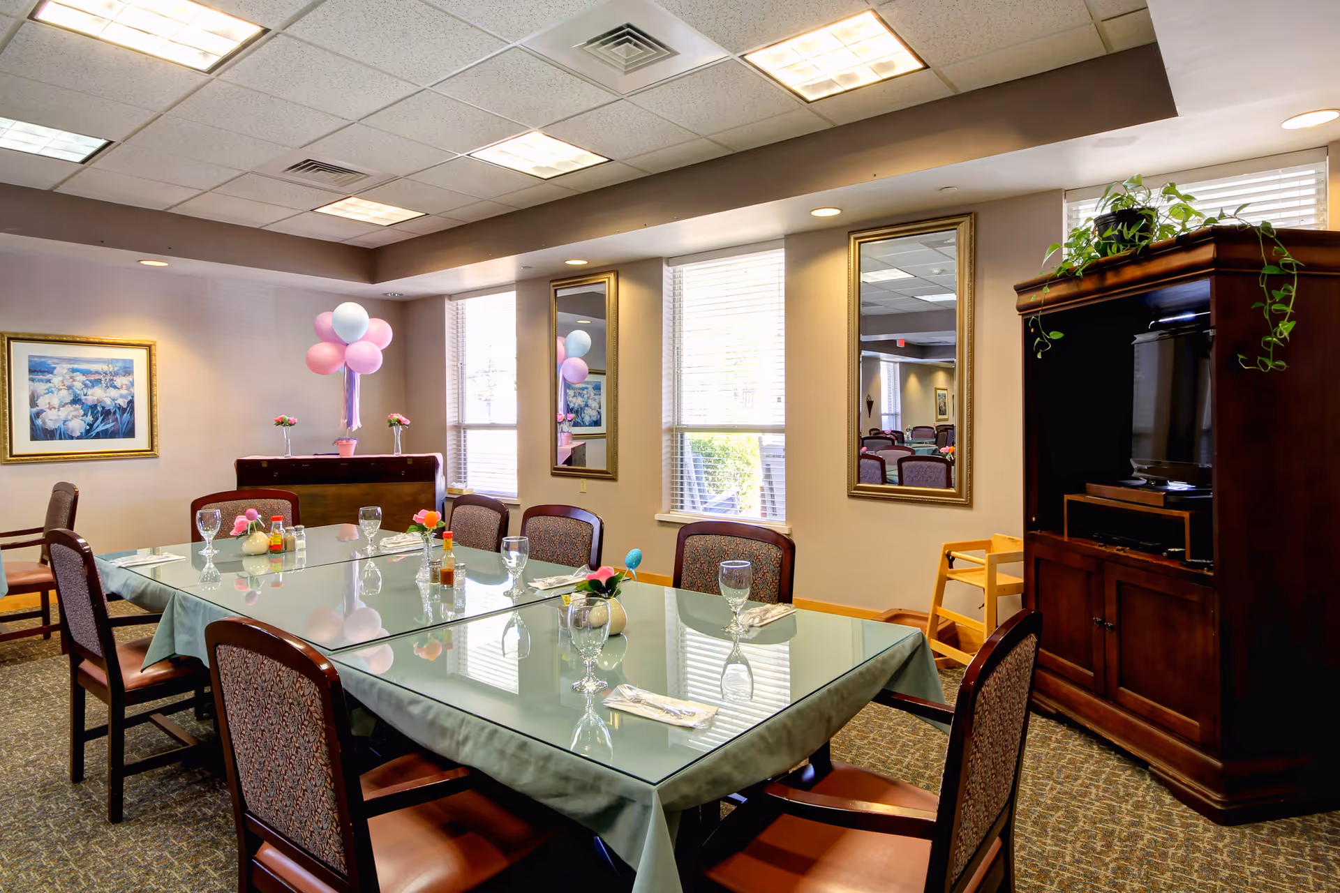 A dining room with a large glass-top table covered by a green tablecloth, surrounded by eight chairs with patterned upholstery. The table is set with glassware, napkins, and small flower arrangements. The room has beige walls with two large windows and two mirrors. There is a wooden cabinet with a TV and a plant on top, and a small wooden high chair nearby. Pink and white balloons are arranged on a piece of furniture against the wall, and a framed floral painting hangs on the left wall.