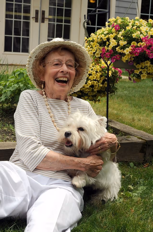 An elderly woman wearing glasses, a white sunhat, and a striped shirt is sitting on the grass outside, smiling and holding a small white dog. Behind her, there are hanging flower baskets with yellow and pink flowers and a building with glass doors and windows.
