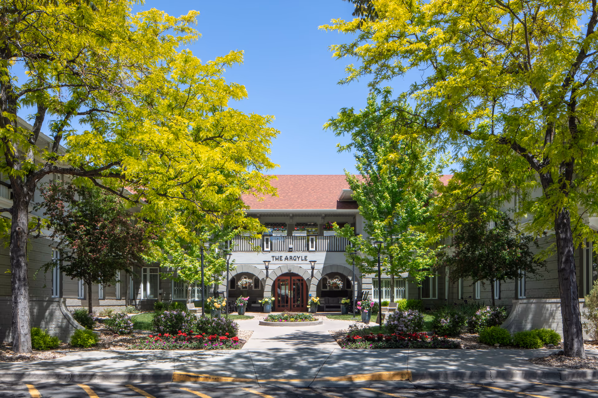 Front exterior view of The Argyle senior living facility with a pathway leading to the entrance, surrounded by green trees and landscaped flower beds under a clear blue sky.