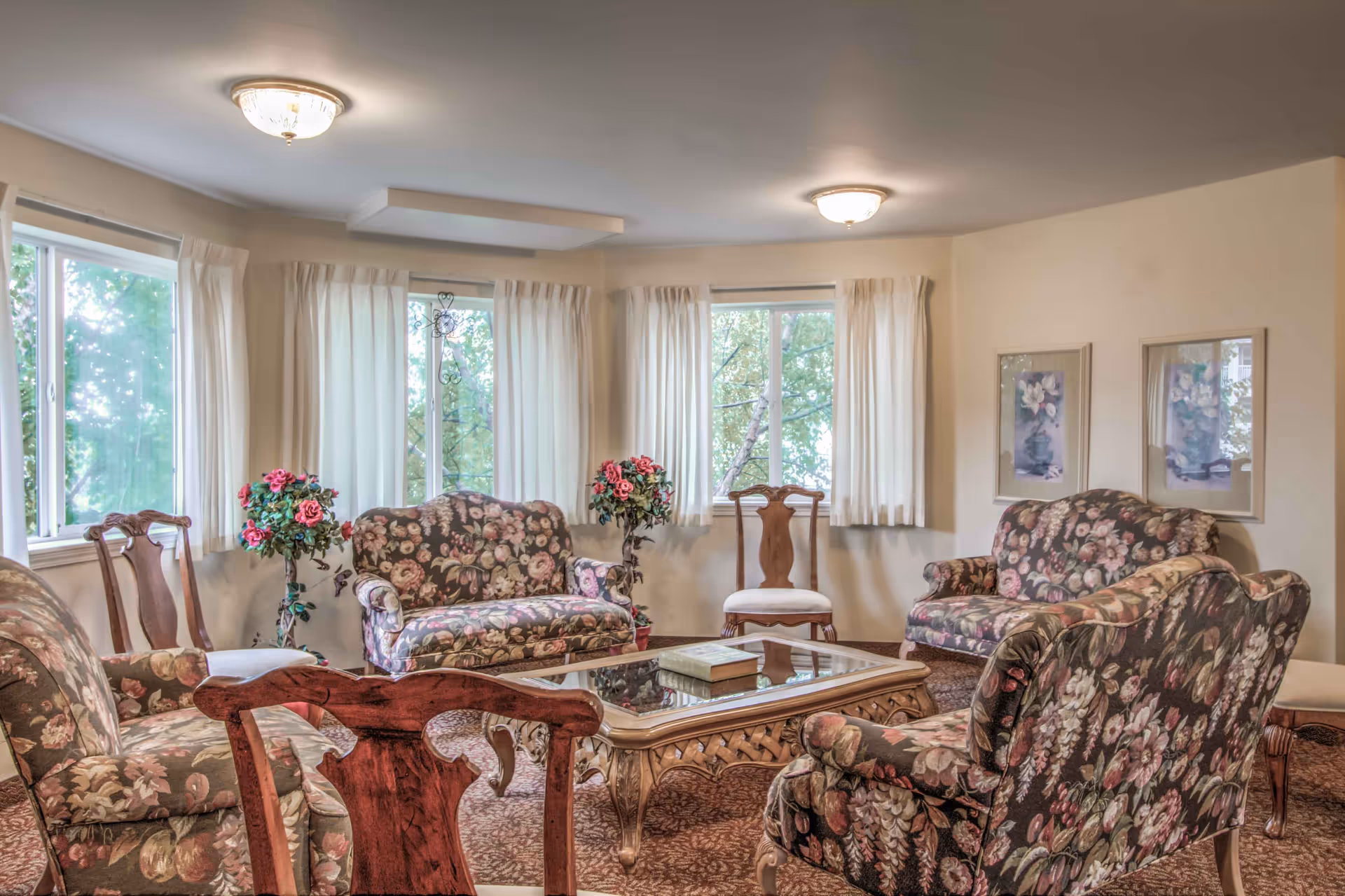 Bright sitting room with floral-upholstered sofas and chairs arranged around a glass-topped coffee table beneath three windows with white curtains.