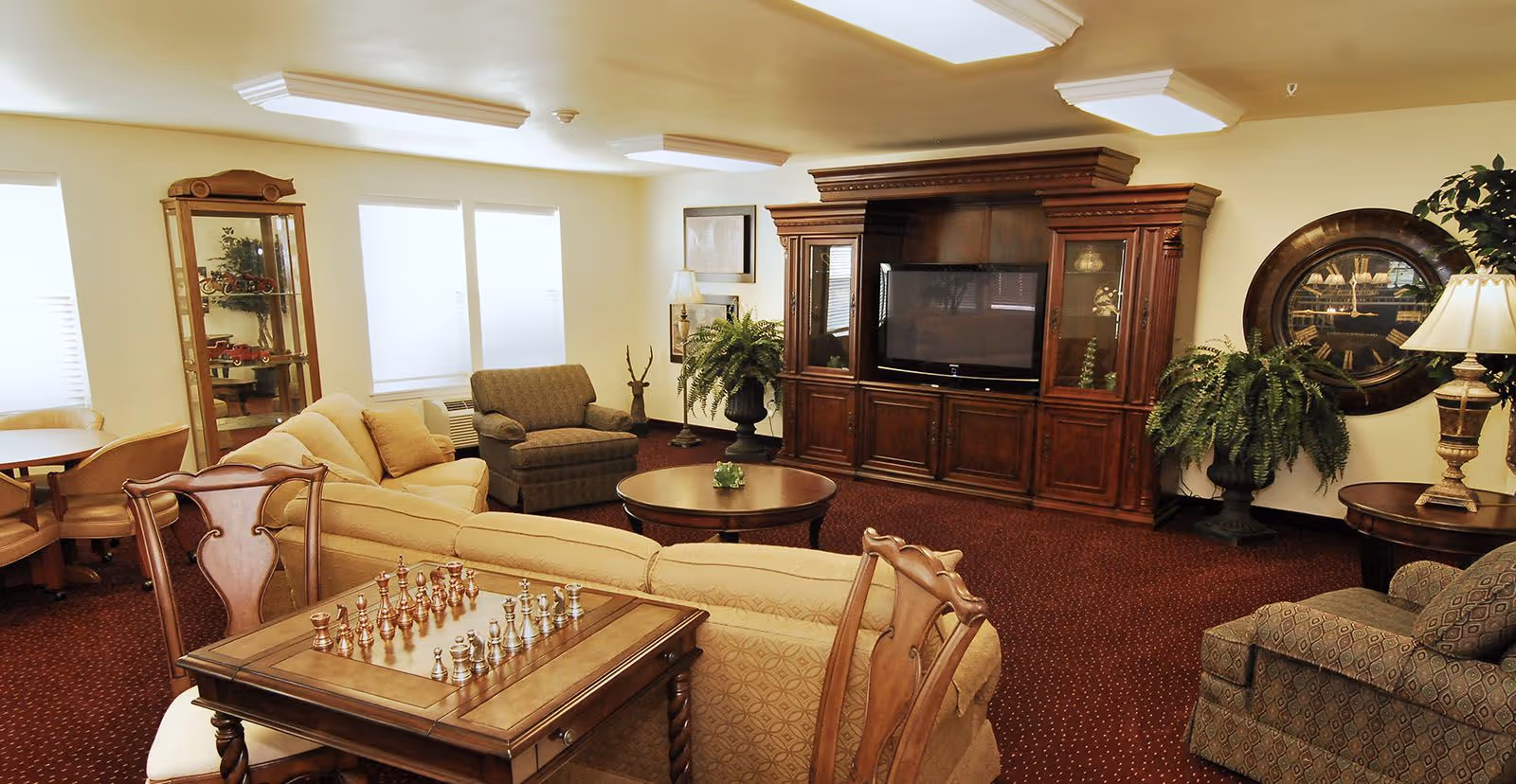 A cozy living room with beige and patterned armchairs, a beige sofa, a wooden entertainment center with a flat-screen TV, a round coffee table, a chess table with chess pieces, and decorative plants. The room has warm lighting and carpeted flooring.
