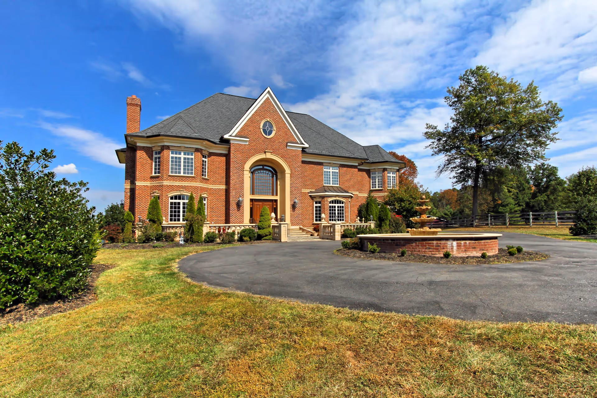 Front exterior view of a large, elegant brick house with multiple windows, a central arched doorway, and a circular driveway with a fountain in the middle. The house is surrounded by well-maintained landscaping and trees under a partly cloudy blue sky.