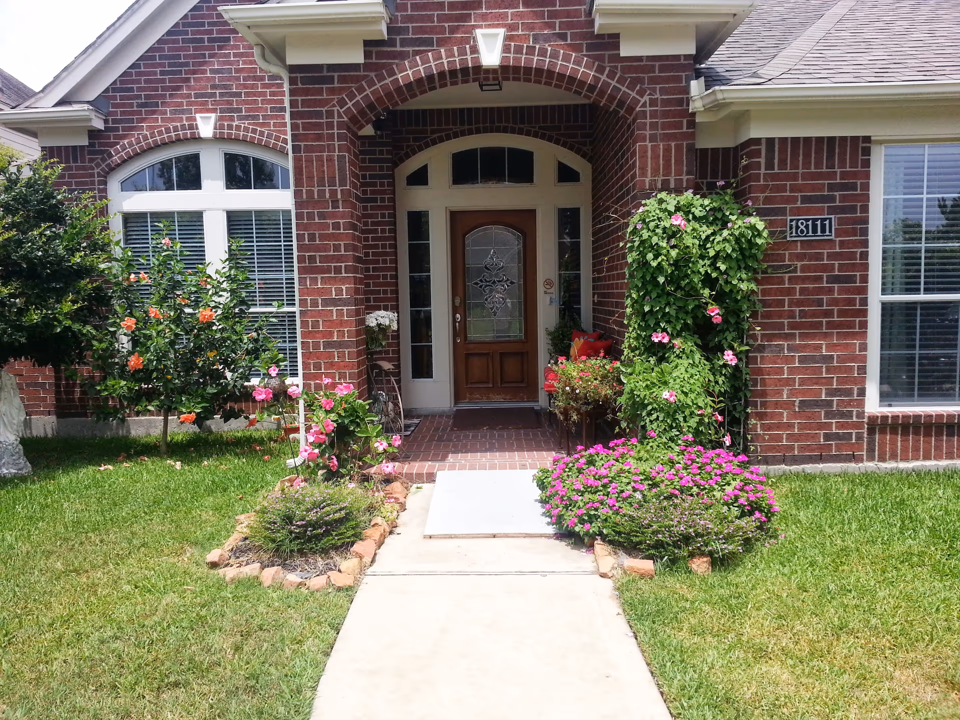 Front entrance of a brick building with a wooden door featuring decorative glass. The entrance is framed by an arched brick design and flanked by windows with white trim. There are flower beds with pink and orange flowers on either side of the concrete walkway leading to the door. The building number 18111 is visible on the right side of the entrance.