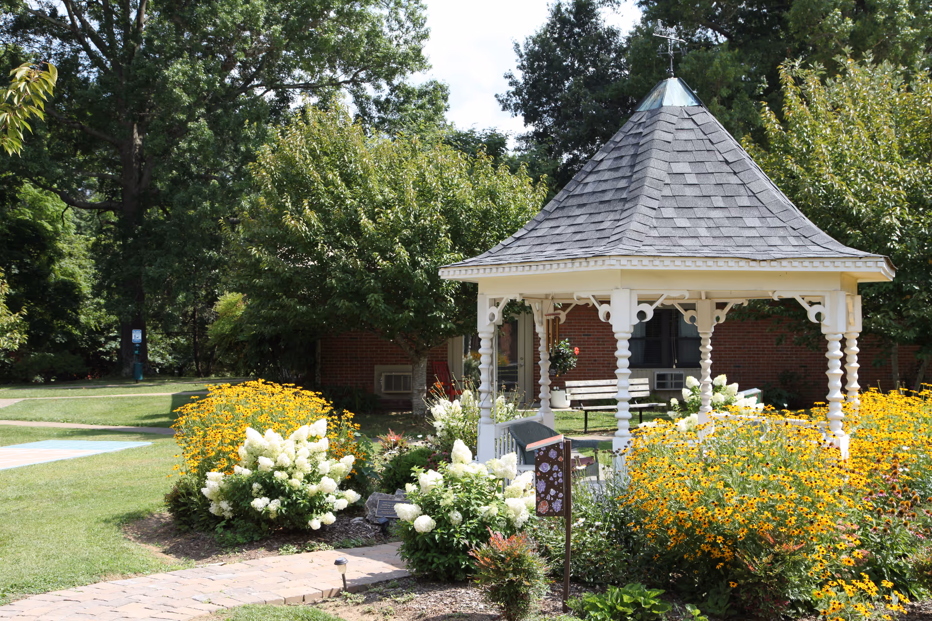 A white gazebo with a shingled roof is surrounded by vibrant yellow and white flowers in a well-maintained garden area. There are trees and a brick building in the background, with a paved walkway leading to the gazebo.