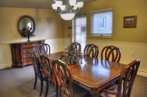 A dining room with a large wooden table surrounded by eight matching wooden chairs. A floral centerpiece is on the table. The room has beige walls with white wainscoting, a window with blinds, a door with glass panes, a wooden sideboard with a vase of flowers, a round mirror above it, and a chandelier hanging from the ceiling.