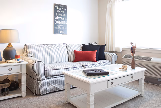 A bright living room with a striped sofa adorned with black and red pillows, a white coffee table with decorative items and books, a side table with a lamp and a cup, a window with white curtains, and a wall decoration with an inspirational quote.