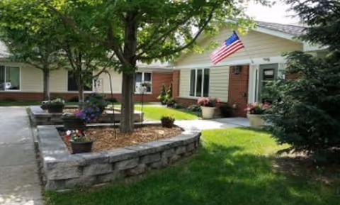 Courtyard in front of a single-story brick and siding building with a raised stone planter, trees and flowers, a manicured lawn, and an American flag by the entrance.