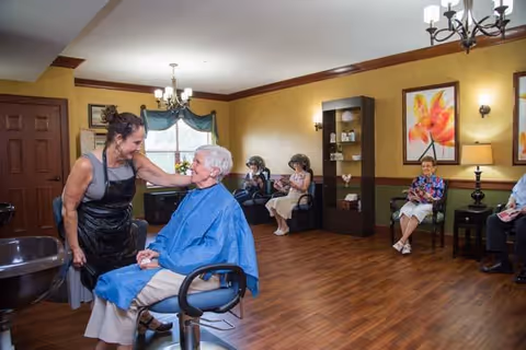 A senior living salon interior with a stylist attending to an elderly woman in a chair while other residents wait in the room.