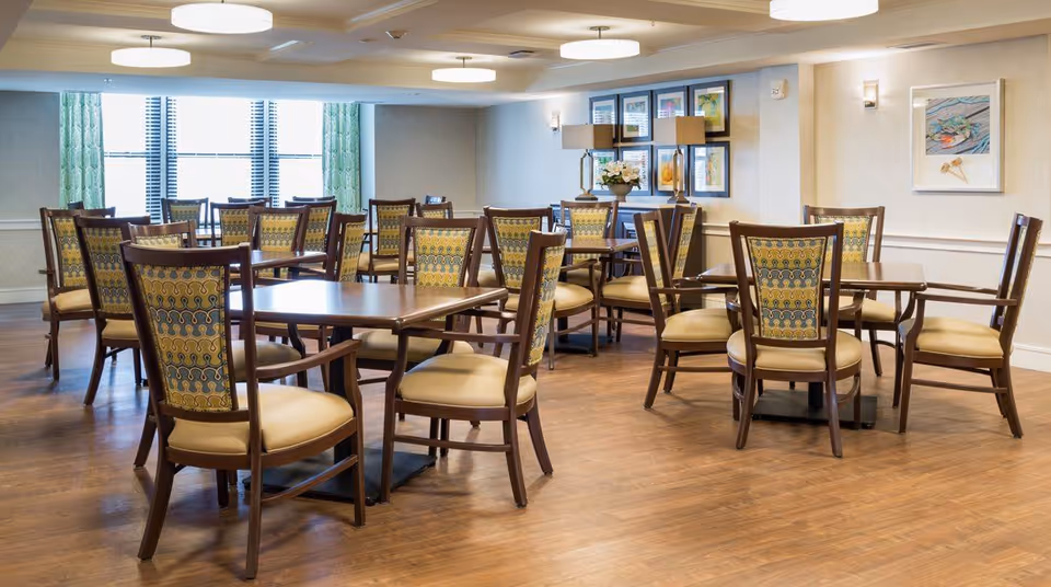 Bright dining room with multiple wooden tables and upholstered chairs on a hardwood floor under ceiling lights.