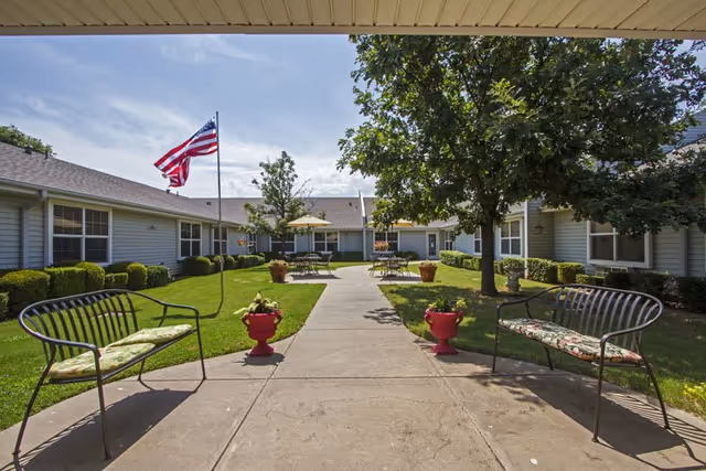 Outdoor courtyard area at Countryside Senior Living of Wichita Falls with a concrete walkway flanked by two metal benches with cushions, potted plants, an American flag on a pole, and tables with umbrellas surrounded by a single-story building and green trees under a partly cloudy sky.