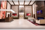 Spacious and modern lobby area with high ceilings, stone accent walls, and contemporary furniture including sofas and chairs arranged around a fireplace and a mounted flat-screen TV. There is a reception desk on the left side and decorative lighting fixtures on the ceiling.